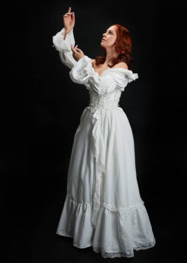 Tam boy portre güzel kadın model, kızıl saçlı, antika beyaz hayalet gelinlik giyiyor. Duruş, karanlık stüdyo arka planında izole bir figür