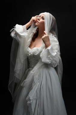 Eski, beyaz, tarihi bir hayalet gelinlik, ruhani bir duvak başörtüsü giymiş güzel bir kadın modelin portresi. Karanlık stüdyo arka planındaki izole bir figürdür..