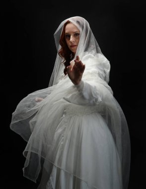 Eski, beyaz, tarihi bir hayalet gelinlik, ruhani bir duvak başörtüsü giymiş güzel bir kadın modelin portresi. Karanlık stüdyo arka planındaki izole bir figürdür..