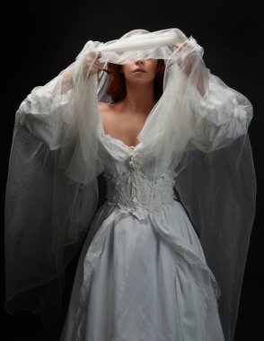 Eski, beyaz, tarihi bir hayalet gelinlik, ruhani bir duvak başörtüsü giymiş güzel bir kadın modelin portresi. Karanlık stüdyo arka planındaki izole bir figürdür..