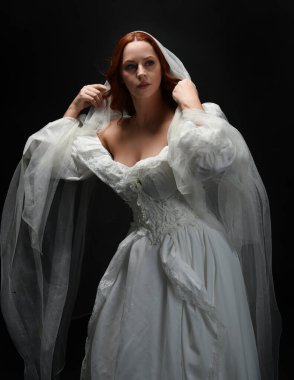 Eski, beyaz, tarihi bir hayalet gelinlik, ruhani bir duvak başörtüsü giymiş güzel bir kadın modelin portresi. Karanlık stüdyo arka planındaki izole bir figürdür..