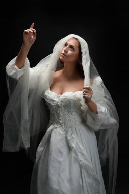 Eski, beyaz, tarihi bir hayalet gelinlik, ruhani bir duvak başörtüsü giymiş güzel bir kadın modelin portresi. Karanlık stüdyo arka planındaki izole bir figürdür..