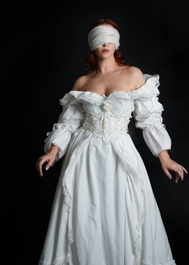 close up portrait beautiful female model wearing antique white historical ghost bride wedding dress gown, a veiled mask blindfold covering face. isolated figure on dark studio background, hands gesturing.