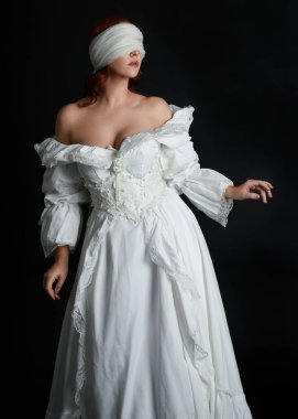 close up portrait beautiful female model wearing antique white historical ghost bride wedding dress gown, a veiled mask blindfold covering face. isolated figure on dark studio background, hands gesturing.