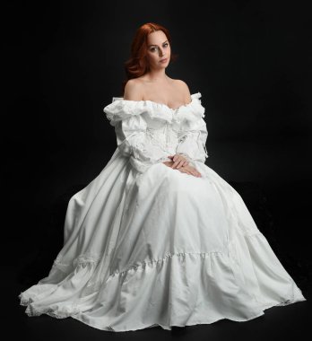 Tam boy portre güzel kadın model antika beyaz hayalet gelinlik giyiyor. Klasik şık oturma pozu, karanlık stüdyo arka planında izole edilmiş bir figür.