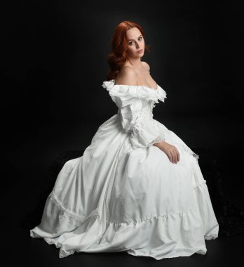 Tam boy portre güzel kadın model antika beyaz hayalet gelinlik giyiyor. Klasik şık oturma pozu, karanlık stüdyo arka planında izole edilmiş bir figür.