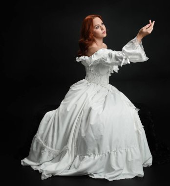 Tam boy portre güzel kadın model antika beyaz hayalet gelinlik giyiyor. Klasik şık oturma pozu, karanlık stüdyo arka planında izole edilmiş bir figür.