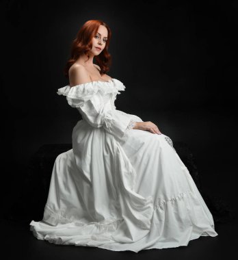 Tam boy portre güzel kadın model antika beyaz hayalet gelinlik giyiyor. Klasik şık oturma pozu, karanlık stüdyo arka planında izole edilmiş bir figür.