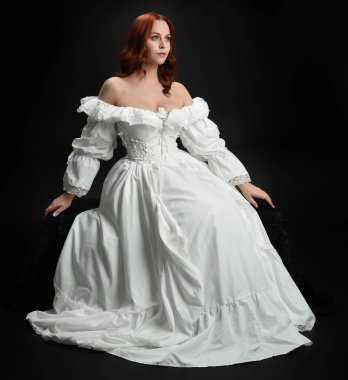 Tam boy portre güzel kadın model antika beyaz hayalet gelinlik giyiyor. Klasik şık oturma pozu, karanlık stüdyo arka planında izole edilmiş bir figür.