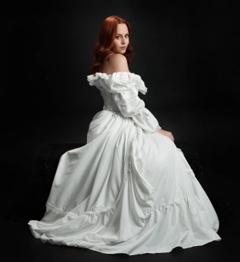 Tam boy portre güzel kadın model antika beyaz hayalet gelinlik giyiyor. Klasik şık oturma pozu, karanlık stüdyo arka planında izole edilmiş bir figür.