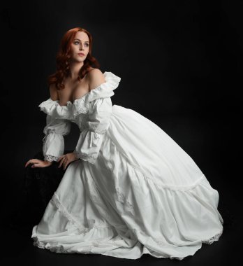 Tam boy portre güzel kadın model antika beyaz hayalet gelinlik giyiyor. Klasik şık oturma pozu, karanlık stüdyo arka planında izole edilmiş bir figür.