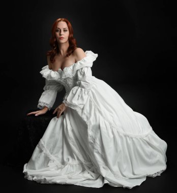 Tam boy portre güzel kadın model antika beyaz hayalet gelinlik giyiyor. Klasik şık oturma pozu, karanlık stüdyo arka planında izole edilmiş bir figür.