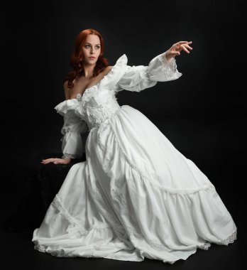 Tam boy portre güzel kadın model antika beyaz hayalet gelinlik giyiyor. Klasik şık oturma pozu, karanlık stüdyo arka planında izole edilmiş bir figür.