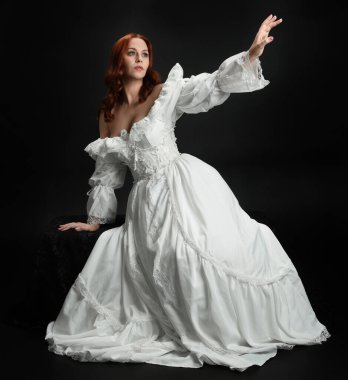 Tam boy portre güzel kadın model antika beyaz hayalet gelinlik giyiyor. Klasik şık oturma pozu, karanlık stüdyo arka planında izole edilmiş bir figür.