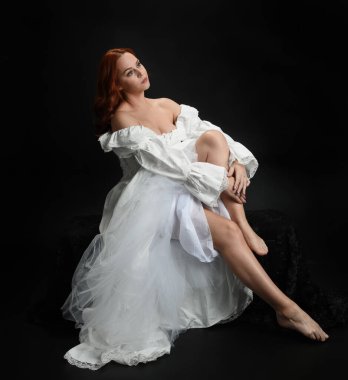 Tam boy portre, kızıl saçlı güzel kadın model, antika beyaz, tarihi hayalet gelinlik giyiyor. Klasik şık oturma pozu, karanlık stüdyo arka planında izole edilmiş bir figür.