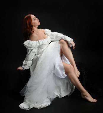 Tam boy portre, kızıl saçlı güzel kadın model, antika beyaz, tarihi hayalet gelinlik giyiyor. Klasik şık oturma pozu, karanlık stüdyo arka planında izole edilmiş bir figür.