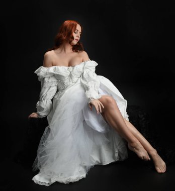 Tam boy portre, kızıl saçlı güzel kadın model, antika beyaz, tarihi hayalet gelinlik giyiyor. Klasik şık oturma pozu, karanlık stüdyo arka planında izole edilmiş bir figür.