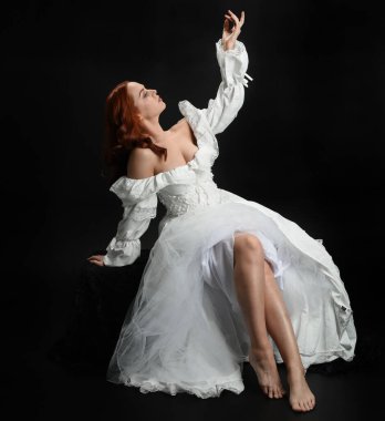 Tam boy portre, kızıl saçlı güzel kadın model, antika beyaz, tarihi hayalet gelinlik giyiyor. Klasik şık oturma pozu, karanlık stüdyo arka planında izole edilmiş bir figür.