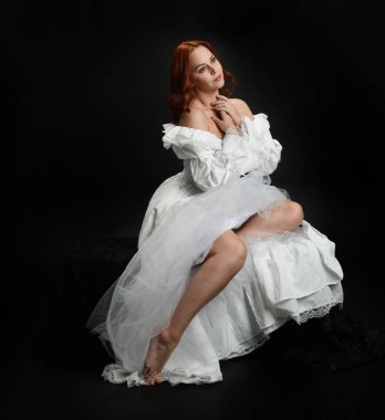 Tam boy portre, kızıl saçlı güzel kadın model, antika beyaz, tarihi hayalet gelinlik giyiyor. Klasik şık oturma pozu, karanlık stüdyo arka planında izole edilmiş bir figür.