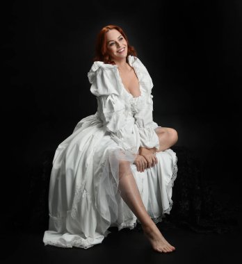 Tam boy portre, kızıl saçlı güzel kadın model, antika beyaz, tarihi hayalet gelinlik giyiyor. Klasik şık oturma pozu, karanlık stüdyo arka planında izole edilmiş bir figür.