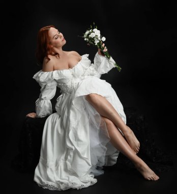 Tam boy portre, kızıl saçlı güzel kadın model, antika beyaz, tarihi hayalet gelinlik giyiyor. Klasik şık oturma pozu, karanlık stüdyo arka planında izole edilmiş bir figür.