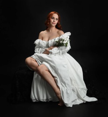 Tam boy portre, kızıl saçlı güzel kadın model, antika beyaz, tarihi hayalet gelinlik giyiyor. Klasik şık oturma pozu, karanlık stüdyo arka planında izole edilmiş bir figür.