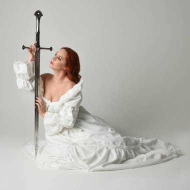 Tam boy portre, tarihi beyaz fantezi gelinlik giyen güzel kadın manken, kılıç tutan peri masalı balosu elbisesi. Stüdyonun arka planında izole bir poz.