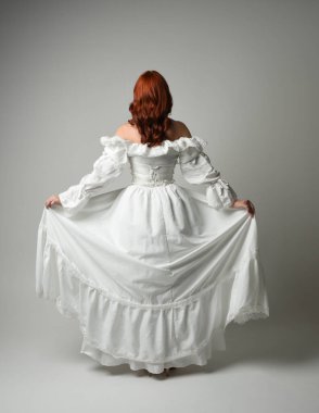 Tam boy, kızıl saçlı, tarihi fantezi gelinlik giyen bir model. Peri masalı balosu elbisesi. Kameradan uzaklaşan poz, beyaz stüdyo arka planında izole bir figür