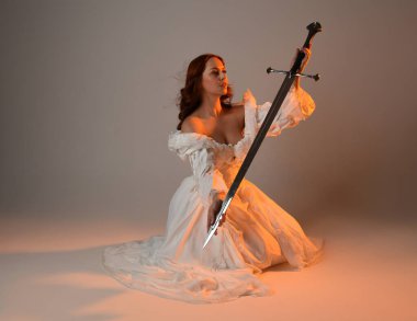 Tam boy portre, tarihi beyaz fantezi gelinlik giyen güzel kadın manken, kılıç tutan peri masalı balosu elbisesi. Stüdyonun arka planında izole bir poz.