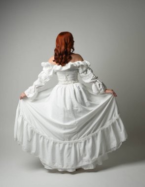 Tam boy, kızıl saçlı, tarihi fantezi gelinlik giyen bir model. Peri masalı balosu elbisesi. Kameradan uzaklaşan poz, beyaz stüdyo arka planında izole bir figür