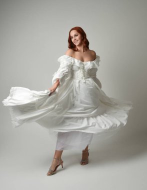 Tam boy, kızıl saçlı, tarihi fantezi gelinlik giyen bir model. Peri masalı balosu elbisesi. Beyaz stüdyo arka planında akan kumaşla izole edilmiş dans figürü