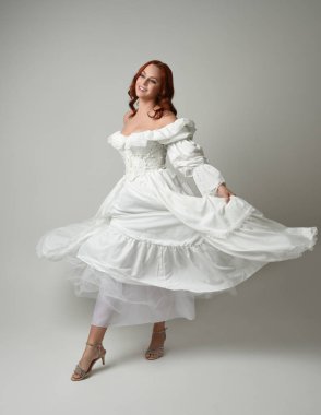 Tam boy, kızıl saçlı, tarihi fantezi gelinlik giyen bir model. Peri masalı balosu elbisesi. Beyaz stüdyo arka planında akan kumaşla izole edilmiş dans figürü