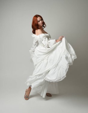 Tam boy, kızıl saçlı, tarihi fantezi gelinlik giyen bir model. Peri masalı balosu elbisesi. Beyaz stüdyo arka planında akan kumaşla izole edilmiş dans figürü