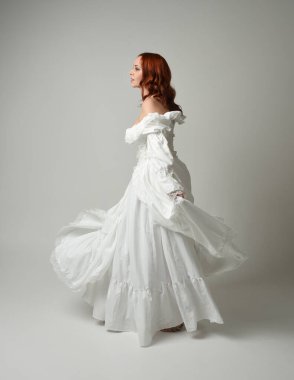 Tam boy, kızıl saçlı, tarihi fantezi gelinlik giyen bir model. Peri masalı balosu elbisesi. Kameradan uzaklaşan poz, beyaz stüdyo arka planında izole bir figür