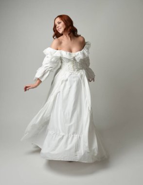 Tam boy, kızıl saçlı, tarihi fantezi gelinlik giyen bir model. Peri masalı balosu elbisesi. Beyaz stüdyo arka planında akan kumaşla izole edilmiş dans figürü