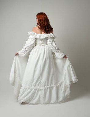 Tam boy, kızıl saçlı, tarihi fantezi gelinlik giyen bir model. Peri masalı balosu elbisesi. Kameradan uzaklaşan poz, beyaz stüdyo arka planında izole bir figür