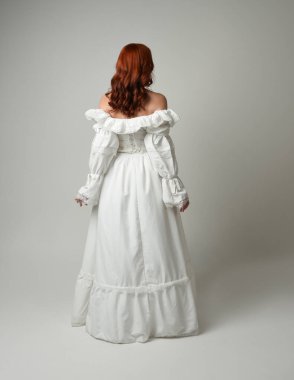 Tam boy, kızıl saçlı, tarihi fantezi gelinlik giyen bir model. Peri masalı balosu elbisesi. Kameradan uzaklaşan poz, beyaz stüdyo arka planında izole bir figür