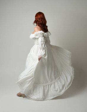 Tam boy, kızıl saçlı, tarihi fantezi gelinlik giyen bir model. Peri masalı balosu elbisesi. Kameradan uzaklaşan poz, beyaz stüdyo arka planında izole bir figür