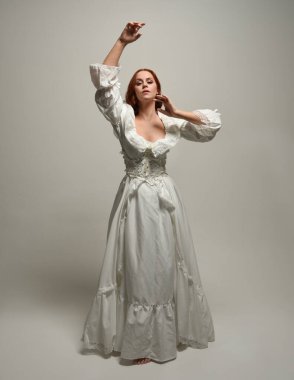 Tam boy, kızıl saçlı, tarihi fantezi gelinlik giyen bir model. Peri masalı balosu elbisesi. Beyaz stüdyo arka planında akan kumaşla izole edilmiş dans figürü