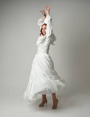 Tam boy, kızıl saçlı, tarihi fantezi gelinlik giyen bir model. Peri masalı balosu elbisesi. Beyaz stüdyo arka planında akan kumaşla izole edilmiş dans figürü