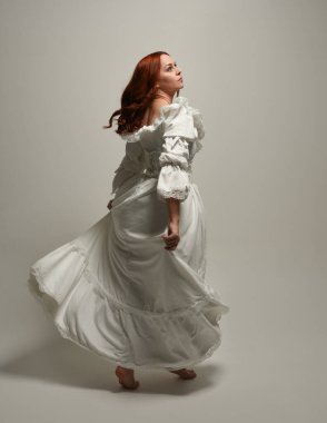 Tam boy, kızıl saçlı, tarihi fantezi gelinlik giyen bir model. Peri masalı balosu elbisesi. Kameradan uzaklaşan poz, beyaz stüdyo arka planında izole bir figür