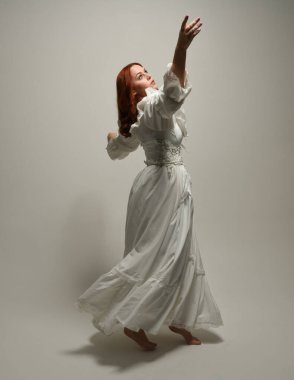 Tam boy, kızıl saçlı, tarihi fantezi gelinlik giyen bir model. Peri masalı balosu elbisesi. Kameradan uzaklaşan poz, beyaz stüdyo arka planında izole bir figür