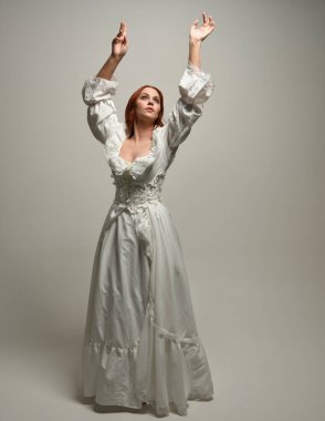 Tam boy, kızıl saçlı, tarihi fantezi gelinlik giyen bir model. Peri masalı balosu elbisesi. Beyaz stüdyo arka planında akan kumaşla izole edilmiş dans figürü