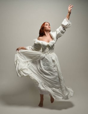 Tam boy, kızıl saçlı, tarihi fantezi gelinlik giyen bir model. Peri masalı balosu elbisesi. Beyaz stüdyo arka planında akan kumaşla izole edilmiş dans figürü