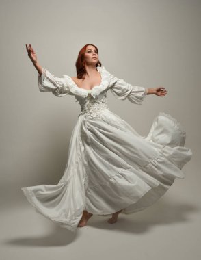 Tam boy, kızıl saçlı, tarihi fantezi gelinlik giyen bir model. Peri masalı balosu elbisesi. Beyaz stüdyo arka planında akan kumaşla izole edilmiş dans figürü