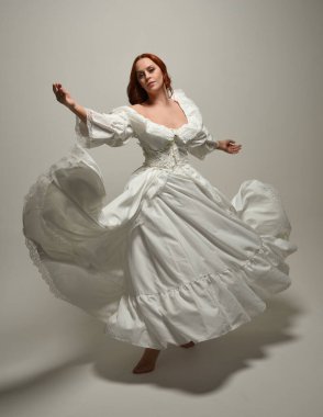 Tam boy, kızıl saçlı, tarihi fantezi gelinlik giyen bir model. Peri masalı balosu elbisesi. Beyaz stüdyo arka planında akan kumaşla izole edilmiş dans figürü