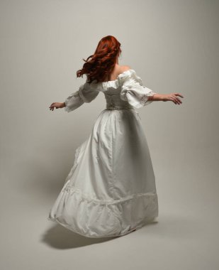 Tam boy, kızıl saçlı, tarihi fantezi gelinlik giyen bir model. Peri masalı balosu elbisesi. Kameradan uzaklaşan poz, beyaz stüdyo arka planında izole bir figür