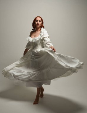 Tam boy, kızıl saçlı, tarihi fantezi gelinlik giyen bir model. Peri masalı balosu elbisesi. Beyaz stüdyo arka planında akan kumaşla izole edilmiş dans figürü