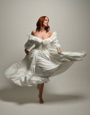 Tam boy, kızıl saçlı, tarihi fantezi gelinlik giyen bir model. Peri masalı balosu elbisesi. Beyaz stüdyo arka planında akan kumaşla izole edilmiş dans figürü