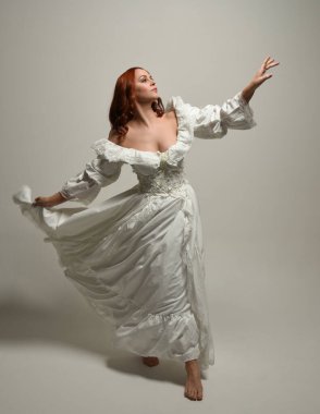 Tam boy, kızıl saçlı, tarihi fantezi gelinlik giyen bir model. Peri masalı balosu elbisesi. Beyaz stüdyo arka planında akan kumaşla izole edilmiş dans figürü